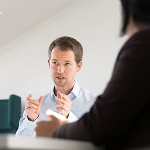 Mann spricht gestikulierend mit zwei anderen Personen in einem Meetingraum und erläutert einen Gedanken.
