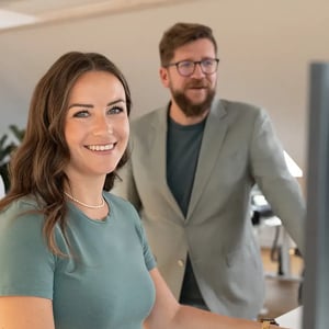 Drei Personen stehen vor einem Bildschirm im Büro, eine Frau in grünem Shirt lächelt direkt in die Kamera.